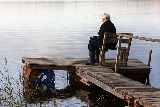 Vanha nainen istuu laiturilla kumisaappaat jalassa ja katsoo maisemaa.