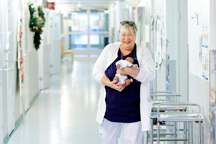 Vaaleatukkainen perushoitaja seisoo sairaalan käytävällä pieni vauva sylissään.